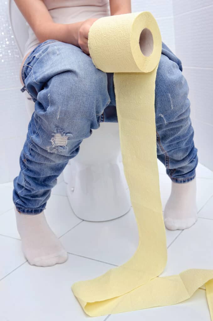 Person sitzt auf einer Toilette und rollt zu viel Toilettenpapier aus.