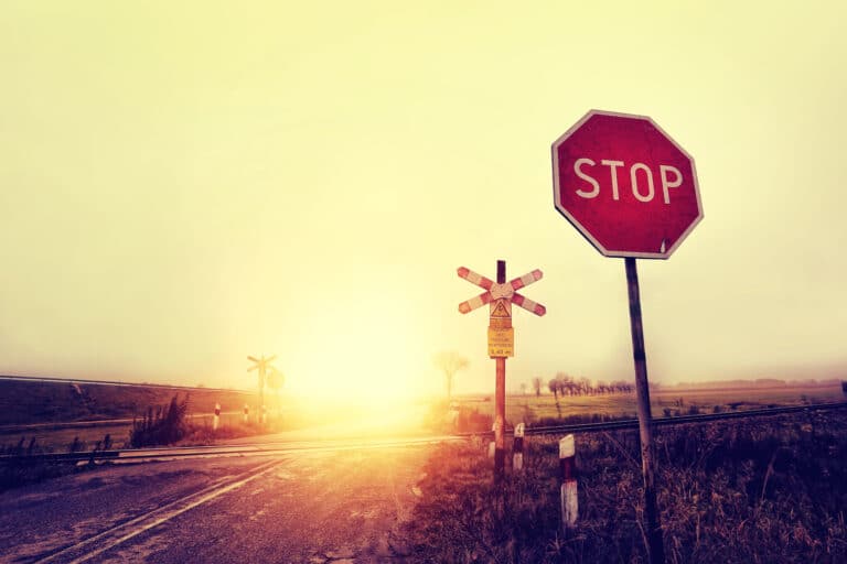 Eine Landstraße kreuzt Bahngleise, die mit einem „STOP“-Schild und Warnschildern für Bahnübergänge gekennzeichnet sind. Im Hintergrund geht die Sonne unter und wirft einen warmen Schein auf die Szene, als ob die Natur selbst wüsste, wo Grenzen gesetzt sind.