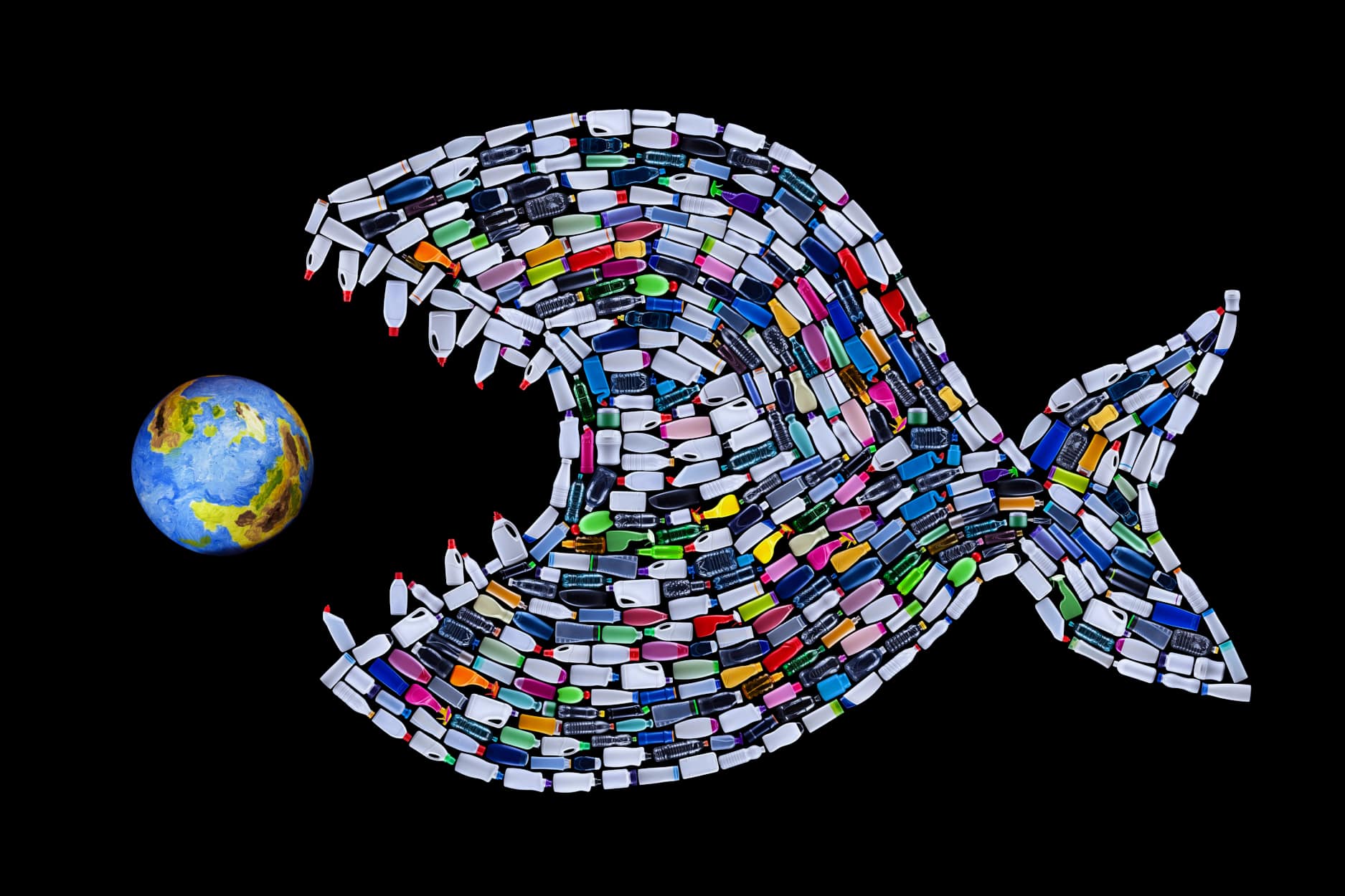 Eine große Fischform aus bunten Plastikflaschen scheint vor einem schwarzen Hintergrund einen kleinen Globus zu jagen und verdeutlicht so die Gefahr von Mikroplastik in unseren Ozeanen.